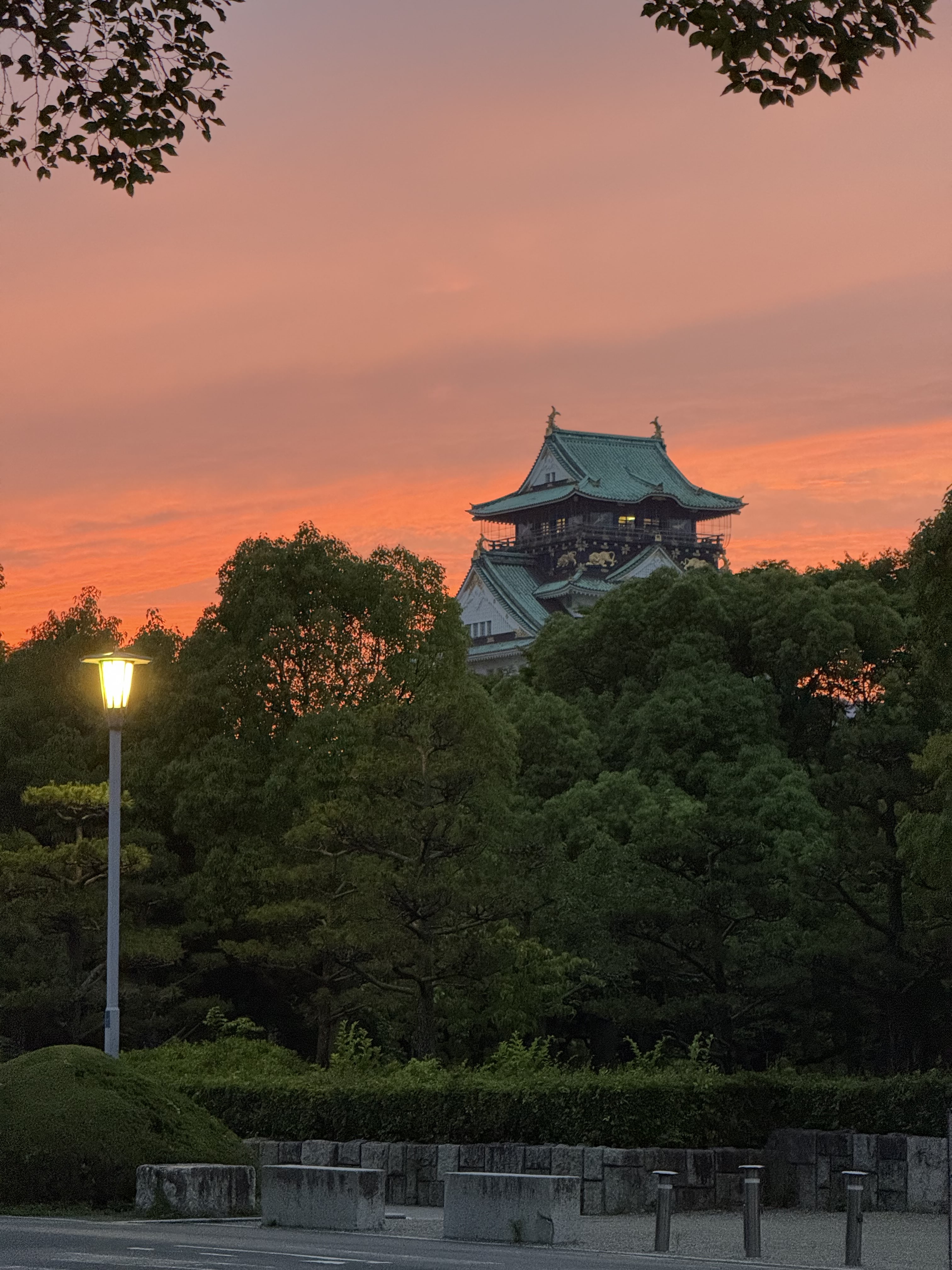 Osaka castle at dusk, surrounded by forrestry with a warm lit streetlamp in view
