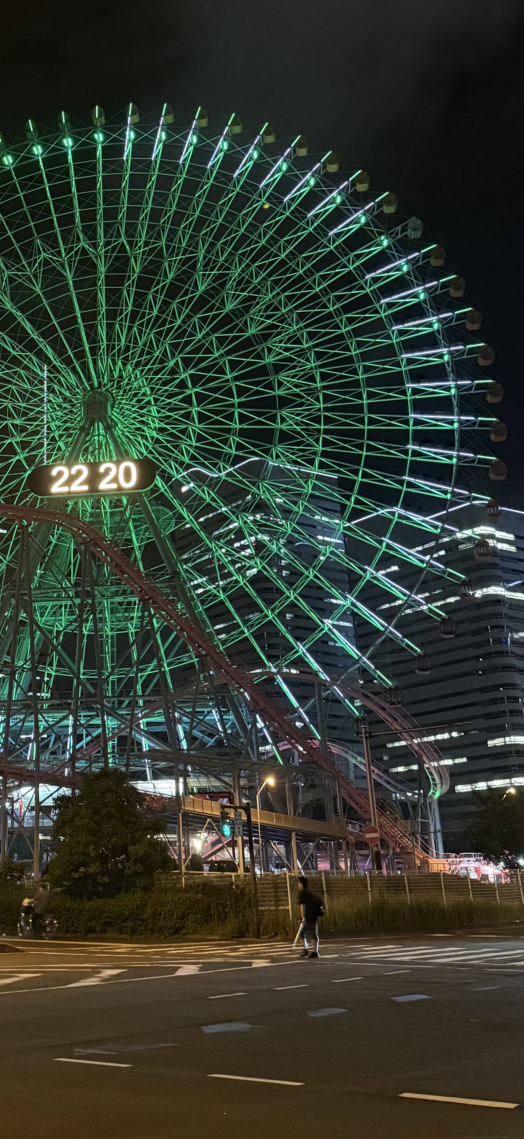 A green lit ferris wheel located in Yokohama, with a man crossing the warm well lit street underneath