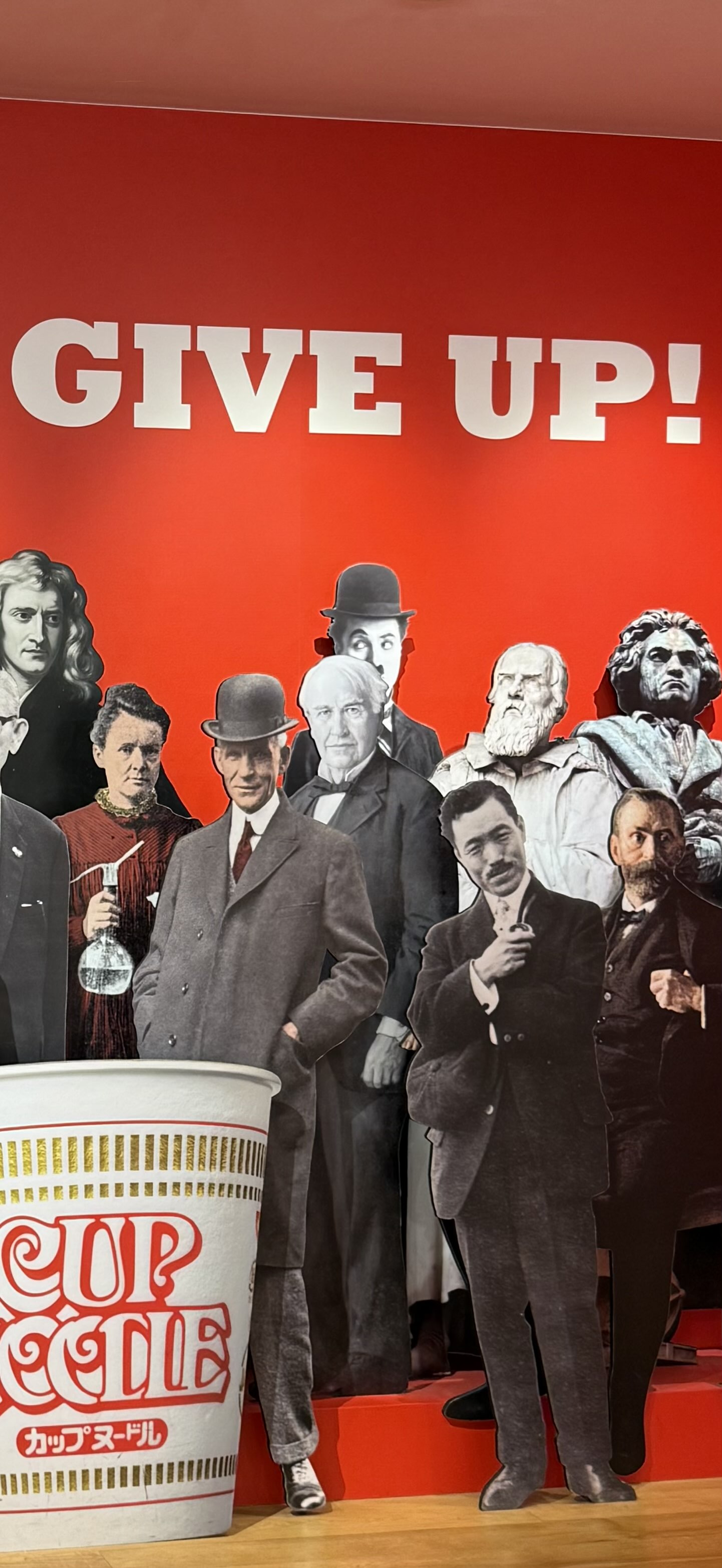 Half of an image wall in the Yokohama Cup Noodle Museum and Factory of historic legends with Never Give Up written across the top